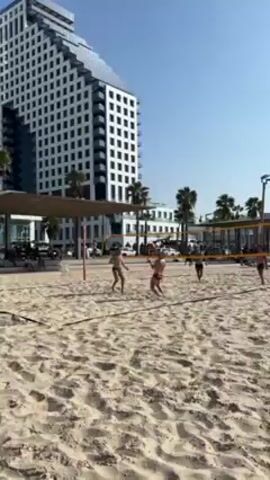 The video shows a beach volleyball game being played on a sandy beach on a clear, sunny day. There are at least six people visible, divided into two teams on either side of a volleyball net. They are wearing casual beach attire, including swimwear and shorts. The players are actively engaged in a volley, moving around to position themselves and hit the ball. In the background, there's a modern, high-rise building with a unique, geometric design, and palm trees dotting the landscape. There are also shaded areas with people relaxing, suggesting it's a public beach. The overall impression is a pleasant, active scene on a sunny beach.