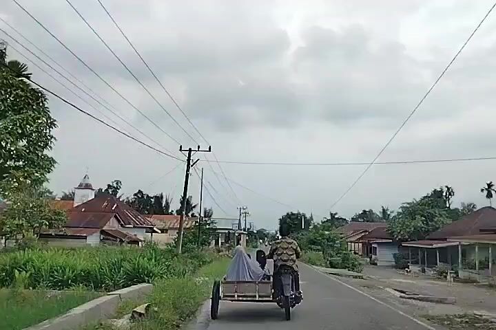 A motorcycle with a sidecar carrying two people travels down a paved road. The motorcycle is being ridden by a person wearing a camouflage shirt. The sidecar appears to be carrying another person covered with a grey tarp. The road is lined with vegetation on both sides, and there are houses and utility poles visible. The sky is overcast.