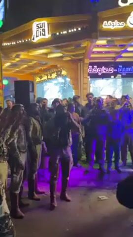 A lively scene unfolds at night, possibly in a Middle Eastern or North African setting. The video captures a group of men, some dressed in what appears to be traditional clothing or military-style uniforms, dancing and singing in a public space. The central figure holds a microphone and appears to be leading the performance. Other individuals in the crowd are watching, filming, and engaging with the performance. The environment is a bustling street with lit storefronts. The lighting is a mix of white and colored lights, including blue and purple hues. The storefronts have signs in Arabic. The overall impression is that of a cultural celebration or a street performance in a vibrant urban setting.