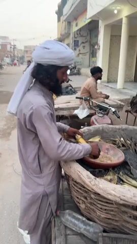 A street vendor in Nasheman Colony, Pakistan, is preparing grilled corn on the cob. The man, with a long beard and wearing a light grey shalwar kameez and a white turban, stands over a rustic cooking setup. He holds a grilled ear of corn and rubs it in a shallow, reddish-brown clay plate filled with a mixture of spices, likely chili powder, salt, and other seasonings.

The cooking station consists of a circular metal basin holding glowing charcoal, which provides heat for grilling the corn. Surrounding this basin is a large, rough, woven basket-like structure, possibly made from thick branches or reeds, holding additional corn cobs, some still partially encased in their husks. A clear plastic water bottle rests on a wooden surface next to the setup.

In the background, a dusty street stretches into the distance. Several buildings line the street, some with visible signs featuring script that appears to be Urdu or Arabic. Other people are visible in the background; a man with a beard sits on a wooden platform to the right, and a child cycles past on a bicycle. The light suggests it is late afternoon or early evening, with a hazy sky indicating a dry, dusty environment typical of the region. The scene captures a moment of daily life and street food preparation.