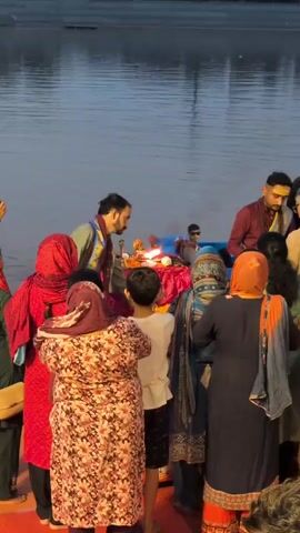 A group of people are gathered near a body of water. In the foreground, several women are facing away from the camera, dressed in traditional clothing including headscarves and patterned dresses. A young boy in a white shirt stands between them. In the center, a man in a red robe and a blue and gold sash is performing a ritual with fire and offerings on a red-covered table. Another man in a maroon outfit stands to the right, also observing. The background shows a large expanse of calm water reflecting the sky. The overall atmosphere suggests a religious ceremony or prayer service taking place outdoors.