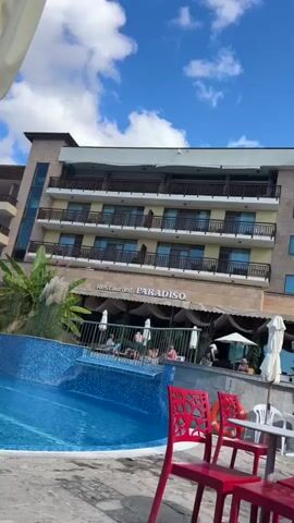The video shows a resort pool area. In the foreground, a white umbrella is partially covering the view of a large blue swimming pool. To the right of the umbrella, two red chairs are visible. In the middle ground, people are seen relaxing by the pool and on lounge chairs. Behind the pool, a multi-story building with balconies and the sign "Restaurant PARADISO" are visible. Lush green plants and tall grasses are planted around the pool. The sky is blue with scattered white clouds. The camera pans slightly across the scene, providing a glimpse of the hotel and the pool area.
