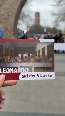 The video shows a recreation of Leonardo Da Vinci's "The Last Supper" in a public space. A group of people are seated at a long table covered with a white tablecloth, resembling the composition of the famous painting. Colored cups and apples are placed on the table in front of each person. The people seem to be posing, mimicking the poses and expressions of the figures in the artwork. The scene is set outdoors, with a brick archway visible in the background. A sign or pamphlet depicting the original artwork is held up in the foreground, reading "Leonardo auf der Strasse," which translates to "Leonardo on the Street". The surrounding environment includes pedestrian traffic, trees, and buildings, indicating an urban setting.