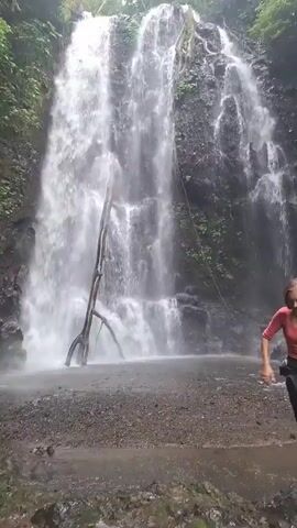 A woman with long, light brown hair is captured in a video in front of a multi-tiered waterfall. She is wearing a reddish-orange top and loose, dark grey pants. Her arms are outstretched and her body is angled as if she is dancing or posing. The waterfall is surrounded by lush green foliage, with a large, weathered log or branch positioned in front of the cascading water on the left side of the frame. The ground in the foreground appears to be composed of dark gravel and dirt. The video appears to be shot outdoors in a natural environment.