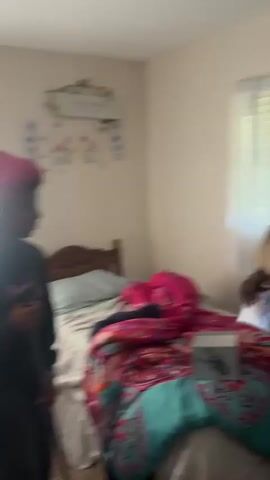 Two young Black women are in a bedroom. One woman wearing a brown wig cap, a tan t-shirt and black shorts is standing by a bed, next to two dark-colored shoe boxes on the bed. The other woman in a red wig cap, a black sweatshirt and black sweatpants is standing on the other side of the bed, holding a Swiffer-type mop. Both women are smiling. The bed has a colorful patterned bedspread. The room is painted white and has a white window with white and purple curtains. There is an art-piece on the wall.