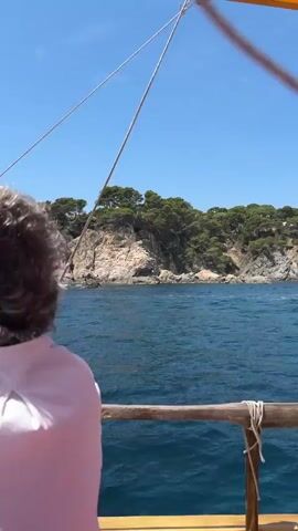 This media file captures a serene day at sea, likely near Palafrugell, Spain. The primary subject is a person, seen from behind, with grey hair and wearing a light-colored, pinstriped shirt and sunglasses. They are seated on a wooden boat, suggesting a leisurely excursion or journey. The boat itself is a key object, with visible wooden railings, ropes, and a red cushion, hinting at a traditional or classic design.

The scene unfolds on a bright, sunny day, with a clear blue sky overhead and calm, deep blue waters stretching out towards a lush, green coastline. The coastline features rocky outcrops and dense vegetation, interspersed with what appear to be houses or structures nestled amongst the trees, indicative of a Mediterranean landscape. Several other boats, including sailboats, are visible in the distance, adding to the maritime setting.

The activity appears to be a boat trip, possibly a sailing excursion, given the presence of masts and rigging visible in some frames. The overall mood is one of tranquility and enjoyment, with the gentle movement of the boat and the scenic coastal views suggesting a peaceful experience. The time of day is clearly daytime, likely late morning or early afternoon, judging by the quality of light and the clear sky. There is no visible text in the media.
