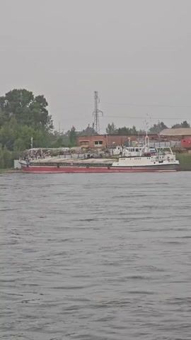 The video shows a wide body of dark water with moderate waves. On the far bank, there is a shoreline with some vegetation and trees. Beyond the vegetation, there are several buildings and industrial structures. Two large smokestacks, one significantly taller than the other, dominate the background, with red and white stripes. There are also other buildings with brick facades and corrugated metal roofs. In the foreground and middle ground on the water, there are two vessels. The first vessel, closer to the left side of the frame, is a long, flat-bottomed barge with a red hull and a white upper structure. It appears to be carrying some cargo or construction materials on its deck. To the right of the barge is a smaller white boat with a red stripe along its waterline, and a cabin structure on its deck. This boat seems to be docked or anchored near the barge. The overall atmosphere is overcast or hazy, suggesting possibly a cloudy or humid day. The camera appears to be moving slowly, likely from another boat on the water, providing a panning view of the industrial waterfront.