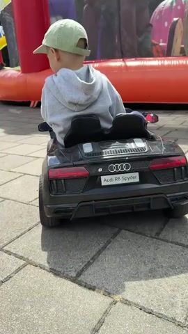 A young boy wearing a gray hooded sweatshirt and a light green baseball cap sits in the driver's seat of a black toy Audi R8 Spyder. He is facing away from the camera, looking towards a large inflatable structure in the background. The inflatable structure appears to be an amusement attraction, with vibrant red, purple, and orange sections visible. The toy car is parked on a paved surface made of interlocking gray pavers. The boy's hands are not visible in the frame. The focus of the video is on the toy car and the boy within it, with the inflatable structure serving as a backdrop. The lighting suggests it is daytime outdoors.