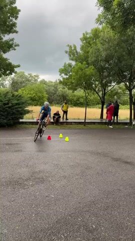 A man in a blue cycling suit and helmet rides a bicycle around three yellow cones on a paved surface. Several people watch under trees in the background; some are holding umbrellas. The weather is overcast and the ground is wet. The cyclist rides away to the right.