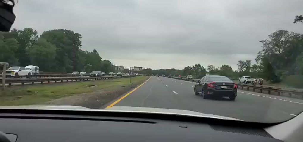 The video is a first-person view from inside a vehicle driving on a highway. The sky is overcast. On the left side of the highway, there are trees and other vehicles, some of which appear to be stopped or moving slowly in traffic. On the right side of the highway, there are also trees and some traffic cones, indicating potential roadwork. The vehicle in front of the camera is a black sedan.