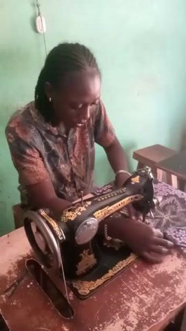 A woman is sitting at a sewing machine. She has dark skin and her hair is braided. She is wearing a short-sleeved button-down shirt with a brown, black, and grey print. The sewing machine is old-fashioned, black with gold decorative accents.  She is using the machine to sew a piece of purple patterned fabric. The sewing machine is on a worn wooden table. The wall behind her is light green.  A light switch and what appears to be a small wooden stool are also visible.