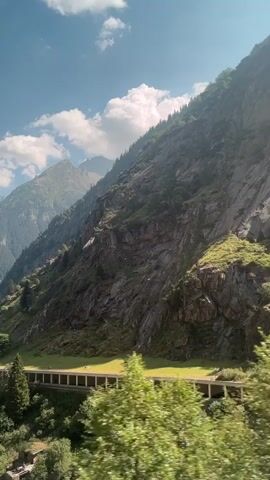 The video displays a scenic view of mountains from a moving vehicle. The mountains are tall and rocky, with patches of green vegetation. There is a clear blue sky with some white clouds. In the foreground, trees are blurred due to the motion of the vehicle. A road with yellow lines is visible, and a tunnel or covered road structure is present on the mountainside. The camera pans and shifts slightly, indicating movement. There are no people or specific interactions visible.