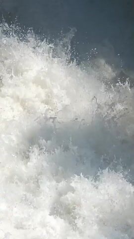 This is a close-up video of churning water, likely a wave crashing or water flowing rapidly over an obstacle. The water is frothy and white with some darker blue or gray areas, suggesting depth or shadows. The motion is turbulent and chaotic, with spray and bubbles visible. There are no people or distinct objects visible, only the dynamic movement of the water itself. The perspective is fixed and focused on the water's surface.