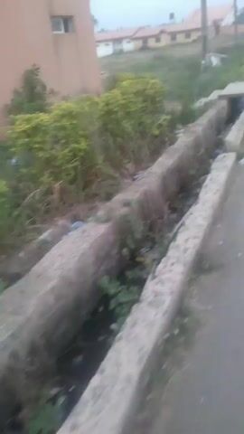 The video shows a concrete drainage ditch running alongside a paved road. The ditch is partially filled with debris and vegetation. On the opposite side of the road from the ditch is a building with a tan exterior and several windows. In the background, there are other buildings and utility poles. A person wearing a yellow shirt can be seen walking away in the distance. The video appears to be taken during the daytime. The scene gives an impression of a residential area with a need for environmental cleanup.