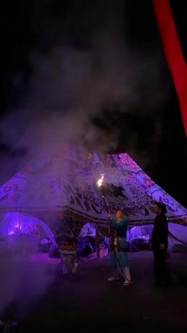 A performer in a colorful costume stands in front of a patterned tent at night, holding a torch that shoots a large flame upwards. Smoke billows around the performer and the tent, illuminated by purple and yellow lights. Another person stands to the right of the performer, watching. A red banner is visible in the upper right corner of the frame.