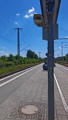 This video shows a view from a train station platform in Mindelheim, Germany. The scene is a bright, sunny day with a clear blue sky dotted with fluffy white clouds. The platform itself is paved with grey tiles, with yellow lines indicating the edge and a metal grate running along the side.

In the foreground, a metal pole with a light fixture and a sign is prominently featured. Further down the platform, a small shelter with blue signs displaying the letter "B" can be seen. Beyond the tracks, there are various buildings, including a larger, older-looking building with multiple windows and a smaller, more modern structure with a sign that reads "Mindelheim." Lush green trees and foliage line the area around the station, and a parking lot with several cars is visible in the distance.

The video pans across the platform, showcasing the train tracks, overhead electrical wires, and the surrounding landscape. The overall impression is one of a quiet, functional train station on a pleasant summer day. There are no people clearly visible in the immediate foreground, though a distant figure can be observed walking further down the platform in one of the frames. The time of day appears to be midday, given the bright sunlight and the shadows cast. The general mood is calm and peaceful.