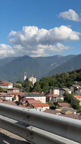 A vertical video shows a small village nestled in a valley, with mountains in the background. The sky is blue with white, fluffy clouds. In the foreground, there is a metal barrier, possibly a guardrail, partially obscuring the view. The village consists of houses with red-tiled roofs, and a prominent church with a bell tower stands in the center of the village, surrounded by trees. The sun appears to be shining, casting shadows and illuminating the scene. The overall impression is of a peaceful, picturesque mountain village on a clear day.