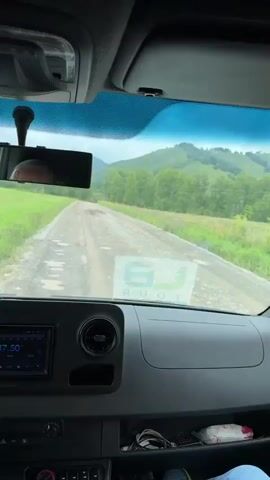 A POV shot from inside a vehicle looking out through the windshield at a dirt road stretching into the distance. The road is flanked by green grassy fields and trees. In the background, rolling hills covered in greenery rise towards a cloudy sky. The dashboard of the vehicle is visible in the foreground, with a media screen displaying 87.50 and a vent. A rearview mirror is also visible.
