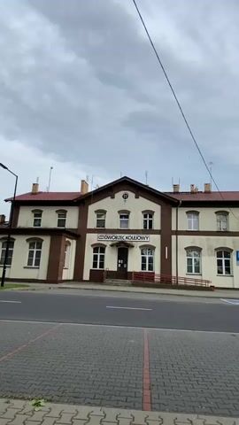 The video opens with a static shot of a two-story building with a cream-colored facade and brown trim, featuring a prominent sign that reads "DWORZEC KOLEJOWY" (Train Station) above its entrance. The building has a brown tiled roof with several small antennas or structures on top. An asphalt road lies directly in front, bordered by a gray paved sidewalk. The sky is overcast and cloudy.

The camera then slowly pans to the right, revealing more of the street and its surroundings. A blue parking sign with a white "P" is visible near the station building. As the pan continues, a silver car is seen parked on the side of the road, next to a modern streetlight. Further to the right, a red brick sidewalk runs parallel to the road, and another building can be glimpsed in the distance amidst green trees. No people are visible, and there is no discernible activity. The cloudy weather persists throughout the video.