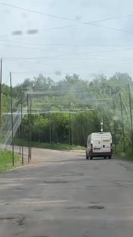 The video is taken from inside a car driving down a rural road in what appears to be Ukraine. The road is cracked and worn. Along one side of the road, a series of tall metal poles are erected, connected by wire and netting to form a tunnel-like structure overhead. The purpose of this structure is not immediately apparent. A white delivery van with Ukrainian lettering and phone numbers on the back is driving away from the camera, further down the road and underneath the netting structure. The background is filled with lush green trees and vegetation. The sky is overcast and gray. The overall atmosphere is somewhat bleak and utilitarian.