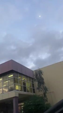 This is a vertical, low-angle shot of a building and the sky. The building appears to be a commercial or institutional structure, with a modern design featuring a prominent corner with a maroon-colored facade and large windows. The windows reflect the sky, showing the interior lights are on, indicating activity within. A tall, slender tree with sparse foliage grows up the side of the building. The sky above is filled with heavy, grey clouds, suggesting overcast or stormy weather. The moon is visible as a small, bright orb in the upper portion of the sky, partially obscured by the clouds. The lighting suggests it's either early morning or late evening, a twilight period. The overall mood is somewhat muted due to the cloudy sky and the architectural style of the building. No text is visible. The location context is Melbourne, Australia.