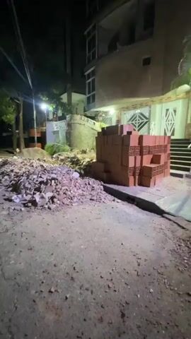 The video shows the aftermath of an apparent explosion or structural collapse at a multi-story building in Maracay, Venezuela. The building, made of brick, has a significant portion of its facade missing on multiple floors, exposing the interior. Debris, including broken bricks and other building materials, is scattered on the ground in front of the structure. A large stack of new red bricks is also visible, suggesting ongoing construction or repair work. The scene is illuminated by artificial lights, indicating it is nighttime. The phrase "DIA OND" is partially visible on a low stone wall to the left of the building, possibly part of a street name or building designation. There are no people visible in the footage. The overall impression is one of destruction and disarray.