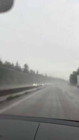 The video is taken from inside a car driving on a wet road during what appears to be a rainstorm. The view is through the windshield, which has raindrops on it, and the windshield wipers can be seen intermittently clearing the view. The road ahead is visible, with lanes marked and other vehicles in the distance, their headlights illuminated. Trees and foliage line the sides of the road, partially obscured by the rain and mist. The overall atmosphere is overcast and gloomy due to the weather conditions. The camera perspective suggests the car is moving forward at a moderate speed.
