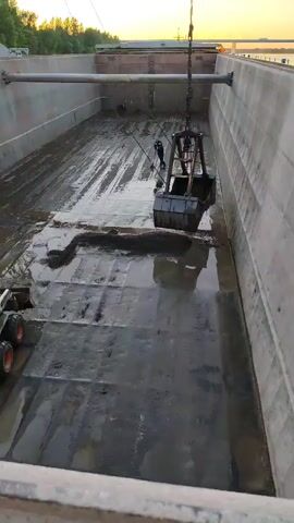 This media file shows an industrial scene within a large, concrete basin. The basin appears to be undergoing cleaning or maintenance, as evidenced by the presence of heavy machinery.

The primary subjects are:

*   **Objects:** A large, mechanical grabber or clamshell bucket is suspended from a crane, actively scooping material from the bottom of the basin. A smaller skid-steer loader, possibly a Bobcat, is positioned on the left side of the basin, with its front attachment lowered, suggesting it is either pushing material or preparing to load it. The basin itself is a prominent object, with concrete walls and a wet, possibly muddy, floor. A large pipe runs horizontally across the upper portion of the basin.
*   **People:** A single person is visible near the grabber, seemingly operating or supervising the machinery.
*   **Scene/Setting:** The overall scene is an outdoor industrial setting. The large concrete basin suggests it could be part of a wastewater treatment plant, a construction site, or a reservoir. The presence of trees and a yellow structure in the background, along with the sky, indicates an exterior location.

The main activity taking place is the removal of sediment or debris from the concrete basin using heavy machinery. The grabber is actively scooping, and the skid-steer loader is positioned to assist in the process.

Notable details include:

*   **Time of Day:** The lighting suggests it is either sunrise or sunset, with a warm, yellowish glow in the sky.
*   **Weather:** The ground is wet, indicating recent rain or the presence of water. The sky appears clear.
*   **Location Cues:** The provided location context is Yaroslavl, Russia. This information, combined with the industrial nature of the scene, points to a facility in that region.
*   **Emotions:** No specific emotions are evident; the scene is purely observational and factual.

There is no visible text in the media file.