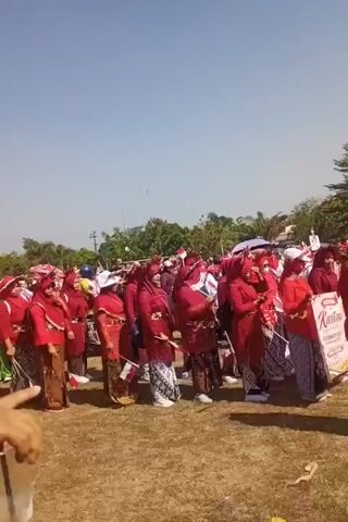A group of people, mostly women, are standing in a line outdoors on a sunny day. They are dressed in traditional Indonesian clothing, predominantly in shades of red and brown. Some of the women are wearing headscarves. A few individuals on the left side of the frame are wearing bright green outfits with red accents. Several people are holding small Indonesian flags. The ground is covered in dry grass, and there are some trees and buildings visible in the background. The sky is clear and blue. The group appears to be participating in a parade or a similar organized event.