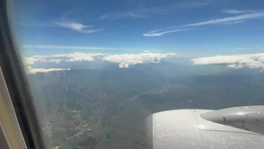 The view from an airplane window shows a mountain range with snow-capped peaks. The mountains are partially obscured by a layer of clouds. Below the mountains, the landscape is green and hilly. In the foreground, the airplane's engine is visible. The sky is blue with some scattered clouds. The overall impression is one of flight and natural beauty.