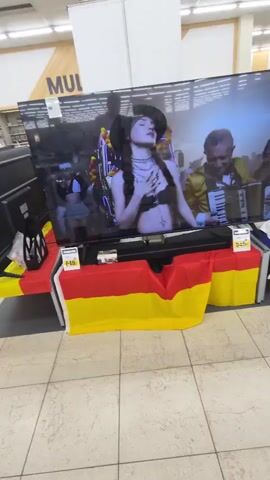 The video shows a large television screen displaying a promotional image of a band. The band members are dressed in elaborate outfits, with one person playing a guitar-like instrument and another playing an accordion. The woman in the center is wearing a revealing outfit. The word "MEXICAN" is prominently displayed on the screen.

The television is on display in a store, likely an electronics retailer. It is set up on a table draped with the German flag, which is a notable detail given the location is Käerjeng, Luxembourg. Price tags are visible on the display, with one reading "649 €" and indicating "PROMOTION" and "LG Téléviseur LED UHD 4K NEW!". Another price tag on the right also reads "649 €". There is also a soundbar placed in front of the TV, with a small promotional brochure for it.

In the background, shelving units with what appear to be other electronics or media are visible, along with store signage that includes the word "MUL". The lighting suggests it is daytime, likely within business hours. The overall scene is typical of a retail environment showcasing electronics. There are reflections on the TV screen, indicating the presence of people and the store environment.