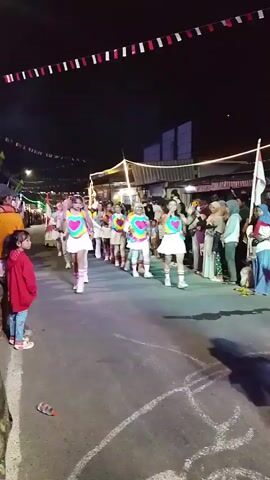 A group of young women in matching colorful outfits with hearts are marching in a parade at night. They are wearing white skirts, colorful tops with heart designs, and pink knee-high socks with white shoes. They are walking down a paved street lined with spectators on both sides. The street is decorated with red and white flags strung across it. The background shows buildings and lights. The overall atmosphere appears festive.