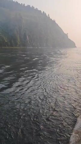 The video opens with a close-up view of a river. The water is dark and choppy, with ripples and small waves visible. On the left side of the screen, a steep, tree-covered mountain rises from the water's edge. The mountain is shrouded in a light mist, and the trees are densely packed. The sky is a pale, hazy orange, suggesting either sunrise or sunset. The camera appears to be moving, as evidenced by the slight blur and the changing perspective of the river and the shoreline. The sound of the moving water is audible, accompanied by a rhythmic, low humming sound. The video continues with the camera panning slightly to the right, revealing more of the river and the mountain range. The mist on the mountain seems to thicken in places. The light remains hazy and muted. The soundscape remains consistent with the initial segment. The video concludes with a further slight pan to the right, showing more of the wide expanse of the river and the forested slopes. The light and mist conditions persist. The sound of the river and the humming continue.