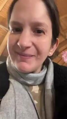 The video shows a woman eating what appears to be a slice of cured meat. She is fair-skinned with brown hair pulled back. She's wearing a gray turtleneck scarf, a dark coat, and a small pearl earring. She holds the thin, marbled slice of meat between her thumb and index finger, brings it to her mouth, bites off a piece, chews, and then looks at the camera with a slight smile. The background appears to be an interior space with a wooden ceiling.  There is indistinct background conversation in a different language. The camera is held close, focusing on her face and the food.