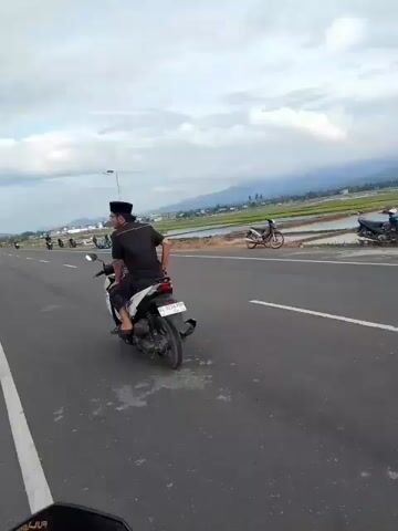 The video shows a road with a motorcyclist in the foreground riding away from the camera. In the distance, other motorcycles are parked on the side of the road, and several people are gathered. The road is paved and appears to be a highway, with a white line marking the lane. On the right side of the road, there are fields, possibly rice paddies, and in the far distance, there are mountains. The sky is cloudy.