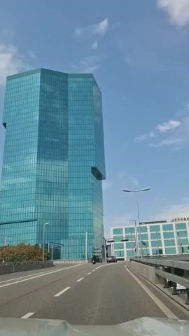 The video shows a view from a car driving on a road. A tall, modern, blue glass skyscraper dominates the left side of the frame. Across the road, there are other modern buildings with blue window frames. Traffic lights are visible, and several cars are on the road. The sky is blue with some white clouds. The sun is shining, creating reflections on the buildings. The car is moving forward, and the perspective shifts slightly as it navigates the road.