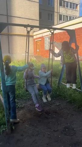 The video shows four young girls on a playground swing set. One girl, wearing a yellow hat and a teal sweatshirt, stands to the left of the swing set, holding onto one of the upright supports. Two girls are seated on a single swing. The girl in the front of the swing is wearing a grey sweatshirt and pants, and the girl behind her is wearing a light blue jacket and pants. Another girl stands to the right of the swing set, on a platform, wearing a patterned jacket and dark pants. The background shows a brick building and a concrete structure. The ground beneath the swing set is dirt and grass. The girls appear to be playing together, with some swinging and others standing.
