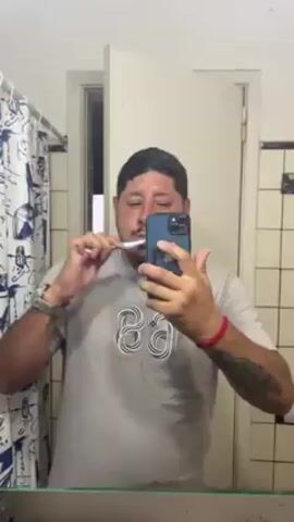 A man is standing in a bathroom, brushing his teeth while holding his smartphone in his right hand to record himself. The man has short dark hair and a trimmed goatee. He is wearing a grey t-shirt with a graphic design, a watch on his left wrist, and a red bracelet on his right wrist. He also has a tattoo on his left forearm. His phone is blue. The bathroom has a white-framed mirror, a blue and white patterned shower curtain, and white and black tiled walls. A white door is partially open behind the man. The environment appears to be lit by artificial light. The faucet is running, and the man is focused on brushing his teeth.