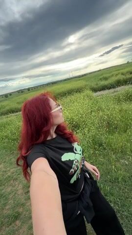 A woman with red hair is taking a selfie in a field of green grass with small yellow flowers. She is wearing sunglasses, a black t-shirt with a green turtle on it, and black pants. She is also wearing a black jacket tied around her waist. The sky is cloudy and overcast. In the distance, there are power lines. The woman is smiling and looking at the camera.