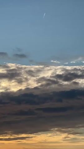 This is a vertical, eye-level shot of a sky with clouds and a contrail from an airplane. The sky is clear and blue in the upper half of the frame. In the middle, there is a band of clouds that are lit from below, appearing white and golden in some areas and dark gray in others. The bottom portion of the frame is filled with darker clouds. A thin white contrail from an airplane is visible in the upper right quadrant of the sky, stretching diagonally across the frame.