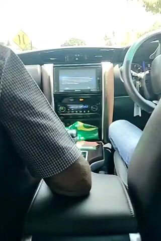The video shows the interior of a car driving on a road. There are two people in the car, the driver and a passenger in the front seat. The driver is holding the steering wheel and is wearing a blue shirt. The passenger is wearing a plaid jacket. The car has a touchscreen display in the center console, and there is a tissue box with green and yellow packaging on the console. The road is lined with trees and buildings, and there are other vehicles on the road, including a truck and motorcycles. The scene appears to be in a tropical or subtropical region.