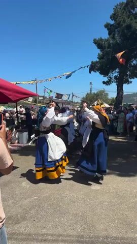 A group of people are dancing in a circle while holding hands. They are wearing traditional clothing. The person in the foreground on the right is wearing a blue dress with a black apron and an orange headscarf. The person in the foreground on the left is wearing a white shirt, a blue and yellow skirt, and a blue headscarf. There are spectators in the background. The event appears to be outdoors under a clear blue sky. There are flags and banners strung across the area, as well as a large tree. There are also market stalls visible in the background. The ground is paved.