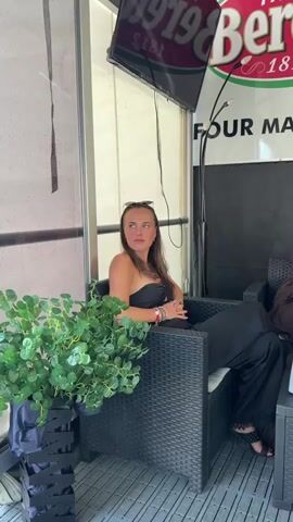 A woman with long brown hair is sitting in a chair. She is wearing a black strapless top and black pants. Sunglasses are perched on top of her head. She is smiling at the camera. A plant is to her left. A TV screen is above her. The environment appears to be outdoors.
