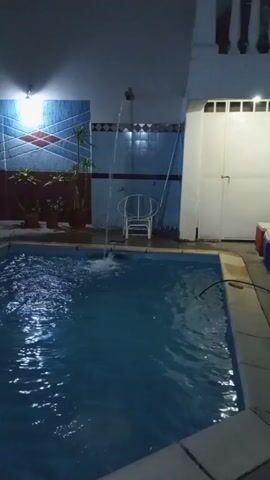 The video shows a rectangular swimming pool at night. The pool is filled with water that reflects the overhead lighting, creating a shimmering effect. A small waterfall feature is visible, pouring water into the pool from a fixture on the wall. The pool is surrounded by tiled flooring. Behind the pool, there is a wall with different colored tiles and potted plants. A white metal chair is placed against the wall. To the right of the pool is a white door. The lighting appears to be artificial. The scene is still, with the only movement being the falling water.