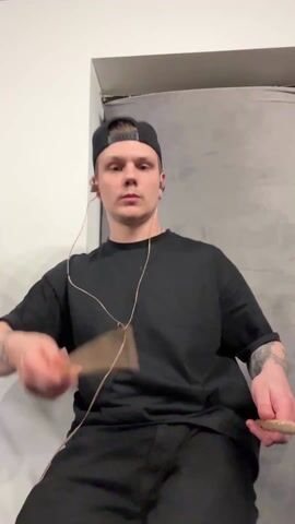 A medium shot depicts a young man sitting down and playing drumsticks in the air. He wears a black t-shirt, black pants, and a black backwards baseball cap. He has tattoos on both forearms. The man is listening to something on earphones, and the wire is visibly running down his shirt. He is twirling and hitting the drumsticks. Behind him is a gray curtain and a white wall. The video is shot at eye level.