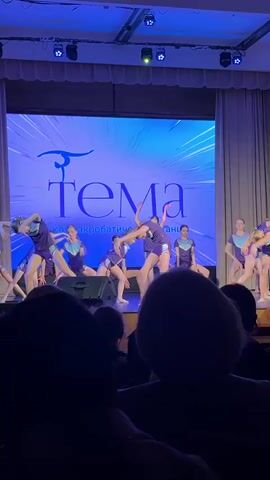A group of young dancers performs on stage in matching purple and teal outfits. They are in sync, bending and swaying to the music. The stage has a large screen behind them displaying the name of their dance club. The audience watches from their seats, their silhouettes visible in the foreground. The stage is lit with bright blue lights.