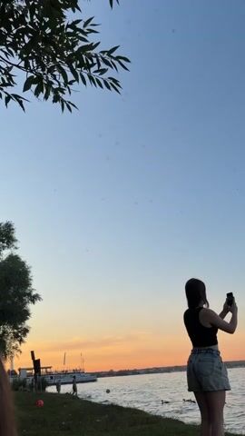 A young woman is standing on grass near a body of water, holding a phone in her hands and taking a photo. She is wearing a black crop top and denim shorts. The sun is setting, casting a warm glow on the sky. There is a boat in the distance and other people on the shore. She then lowers her phone and walks away.