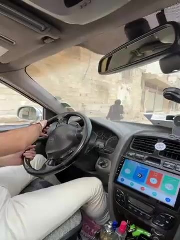 The camera is positioned inside a car, facing forward from the driver's side. A man is seen driving, his hands on the steering wheel and his arm resting on the center console. He is wearing a white watch and light-colored pants. The car has a modern infotainment system with a touch screen displaying a colorful interface. The interior of the car is gray. Through the windshield, a view of a narrow street with aged buildings is visible. There are also people walking on the street. The overall environment appears to be a residential neighborhood.