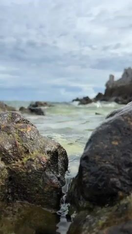 This is a short, low-angle video clip showcasing a rocky coastline with waves gently lapping at the shore. The foreground is dominated by two large, wet rocks, one on the left covered in patches of green and red algae, and a darker, smoother rock on the right. A small trickle of water runs down between these rocks, leading to a pool of water at their base.

In the mid-ground, the shallow, greenish water is visible, with small waves breaking and creating a foamy surface. Beyond the water, a rugged, rocky outcrop rises from the sea, silhouetted against a cloudy sky. The sky itself is filled with grey and white clouds, suggesting an overcast or partly cloudy day.

The time of day appears to be daytime, possibly late morning or early afternoon, given the brightness of the scene. The overall mood is serene and natural, focusing on the textures and movement of the coastal environment. The location is identified as Tambon Phru Nai, Thailand, which is a coastal area known for its natural beauty. There is no visible text in the video.