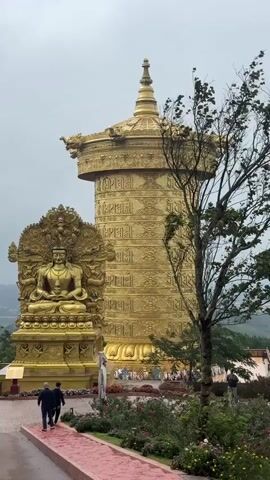The video shows a gold Buddhist prayer wheel and statue in what appears to be a garden setting. Two people are walking away from the camera on a red path. The prayer wheel is large and ornate, with intricate carvings and Buddhist scripture. The statue is also gold and depicts a seated figure, likely Buddha. The environment appears to be overcast and somewhat damp, with greenery visible in the background. The video is shot from a medium distance, providing a clear view of the main subjects.